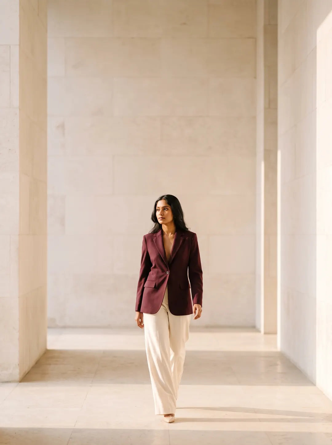 Woman in a wine blazer and cream trousers walking through a sunlit stone corridor